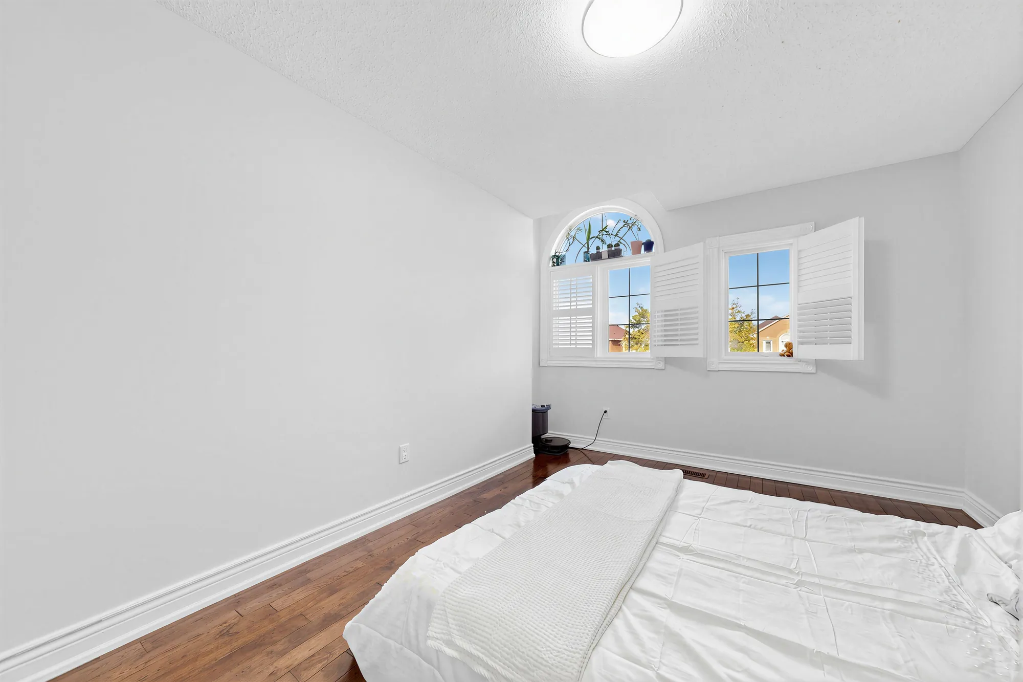 A front bedroom: an arched window and a second window with shutters, hardwood floors, a bed in the foreground