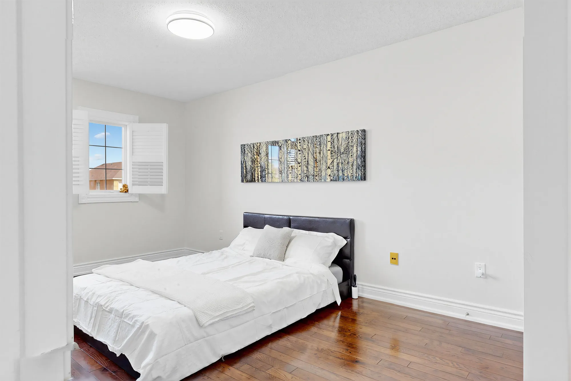 The same bedroom: a bed with a dark headboard below a long print, a window with shutters at the left