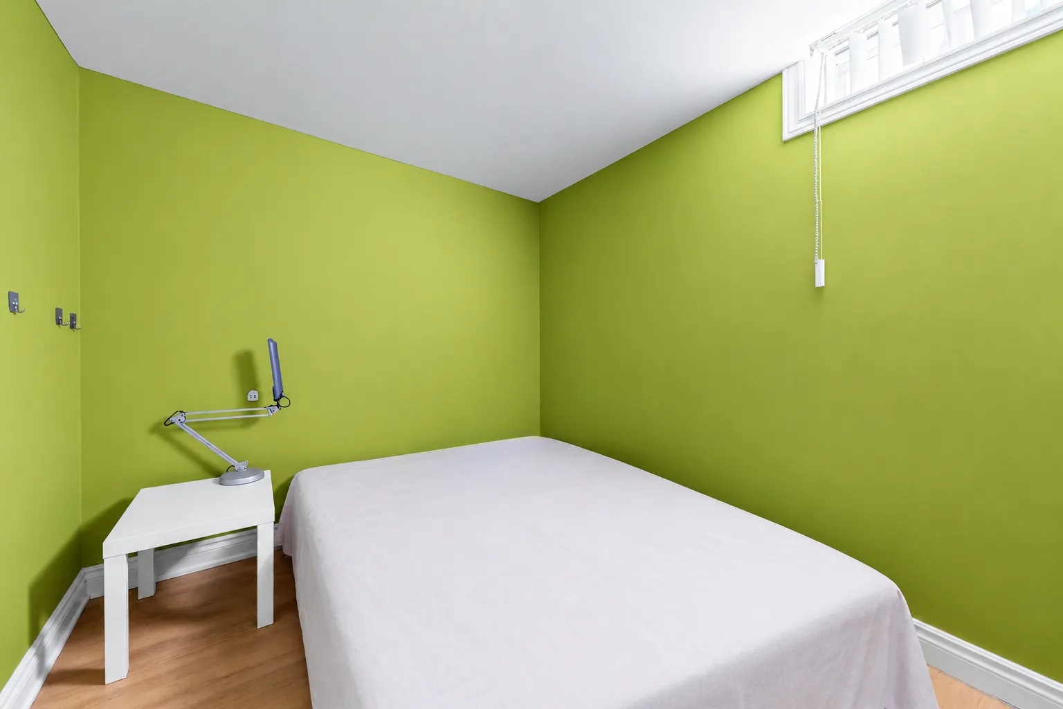 The lower-floor bedroom shown cleared: green walls, a window high on the wall, a bed made up in white, a small white table with a lamp