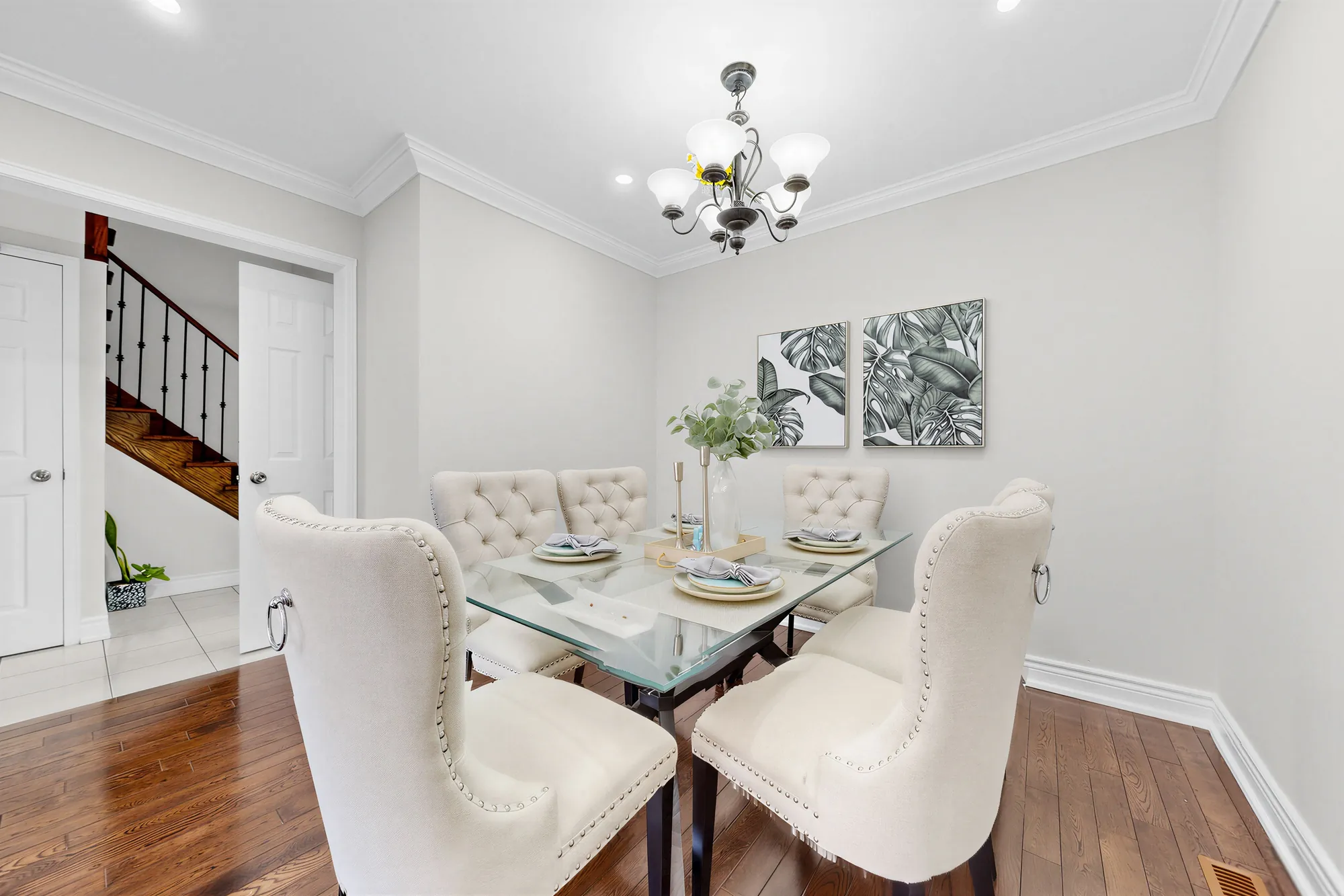 The dining room: a glass table with upholstered chairs under a chandelier, crown moulding, the stair through the doorway at the left