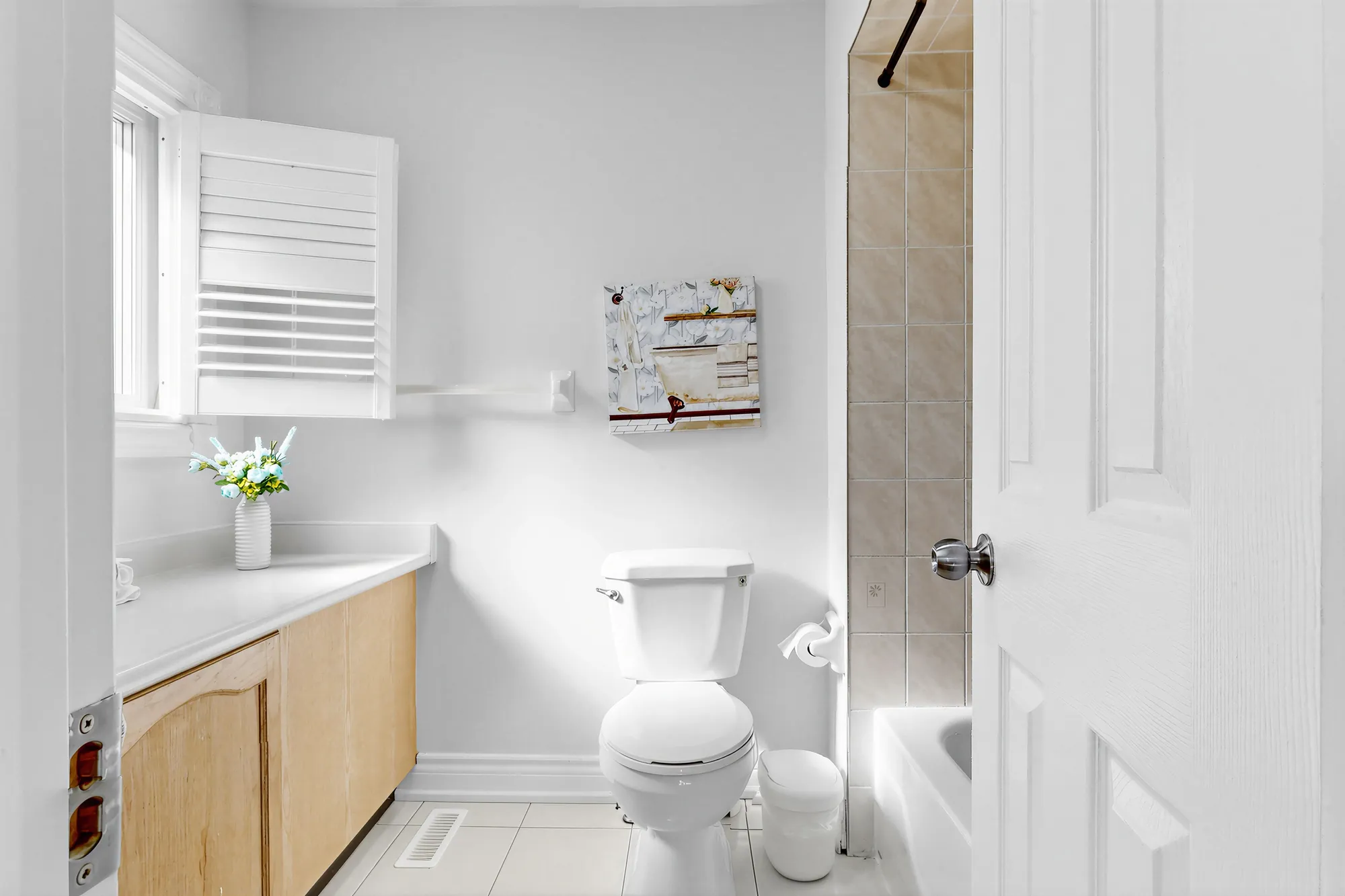 The primary bathroom: a maple vanity with a white top, the toilet, a window with shutters, the tiled tub surround at the right