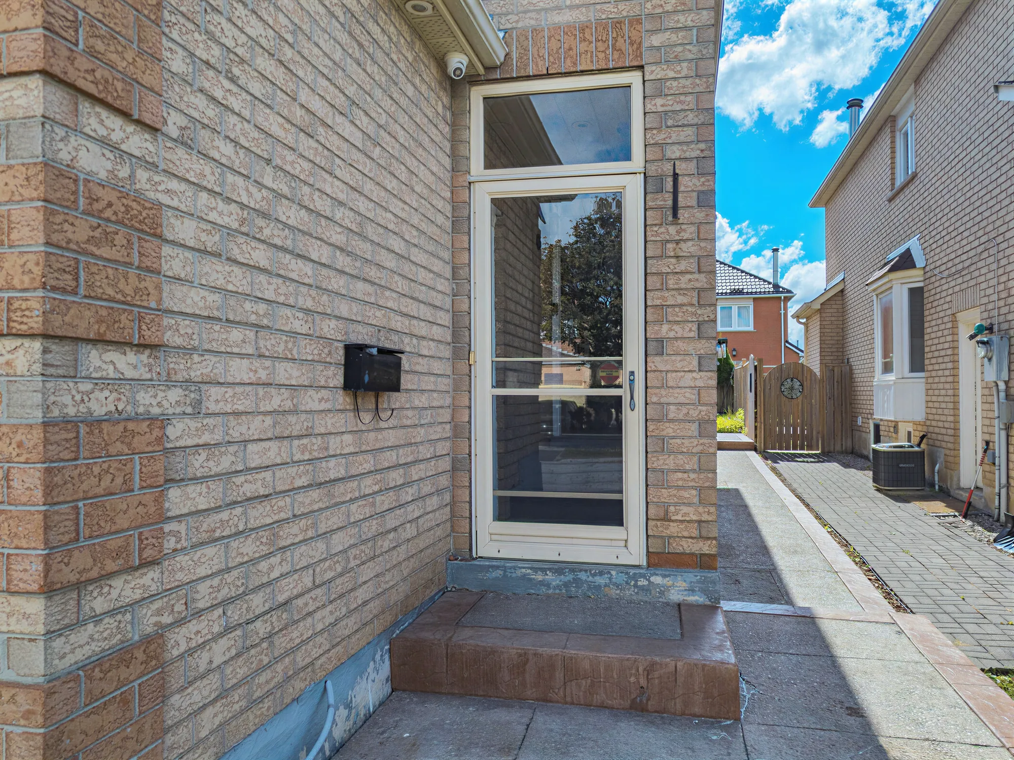 The door into the enclosed porch: a glass storm door in the brick wall, a mailbox, the step, the paved side path beyond