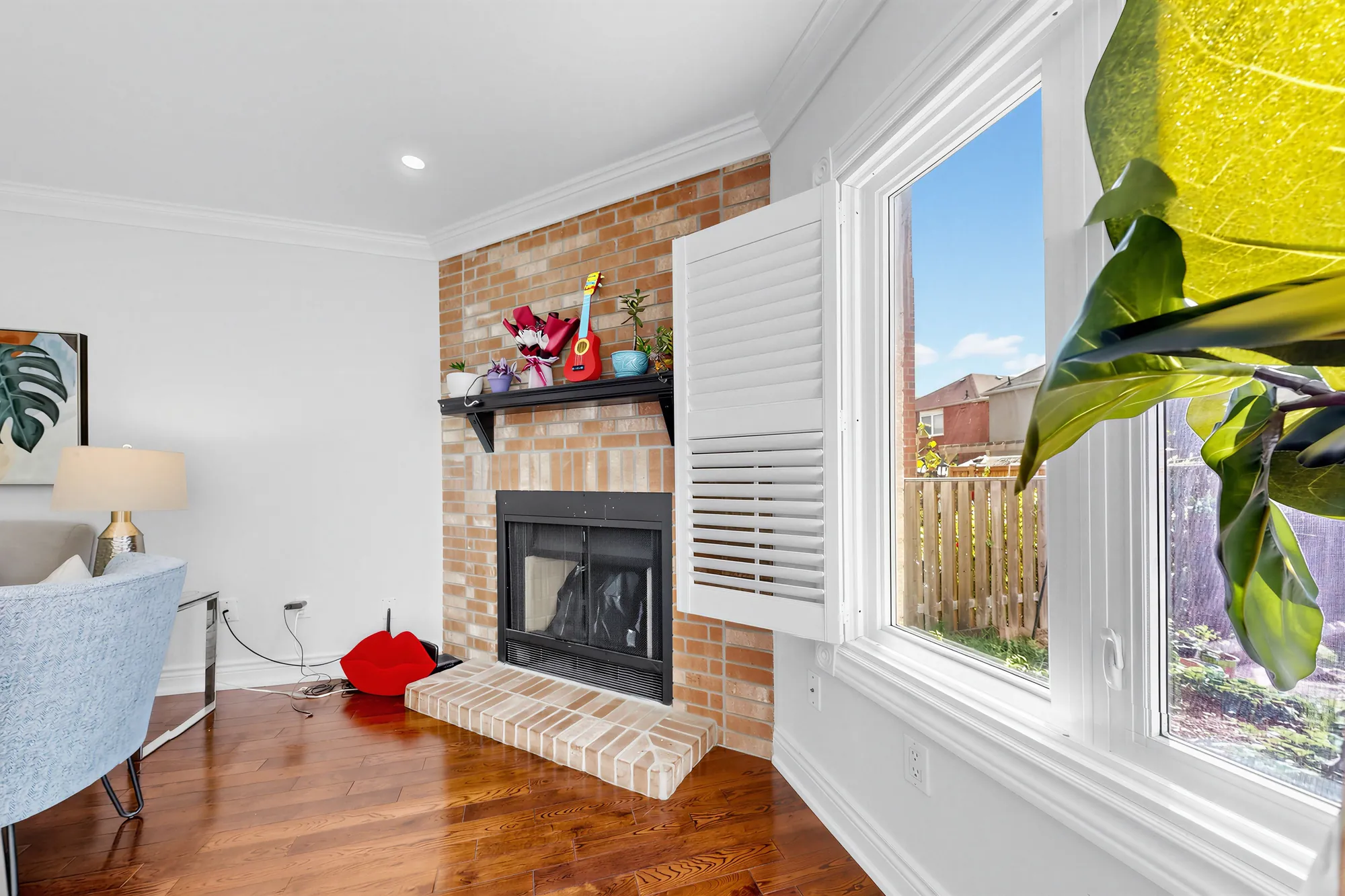 The brick fireplace in the corner of the living room, a window with white shutters beside it onto the back yard