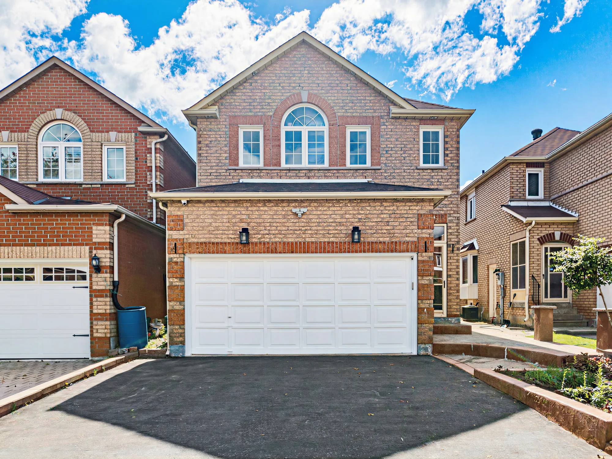 The front of the house by day: two storeys of orange-red brick under a hip roof, an arched window over the white double garage door, a lantern either side of the garage, the asphalt drive