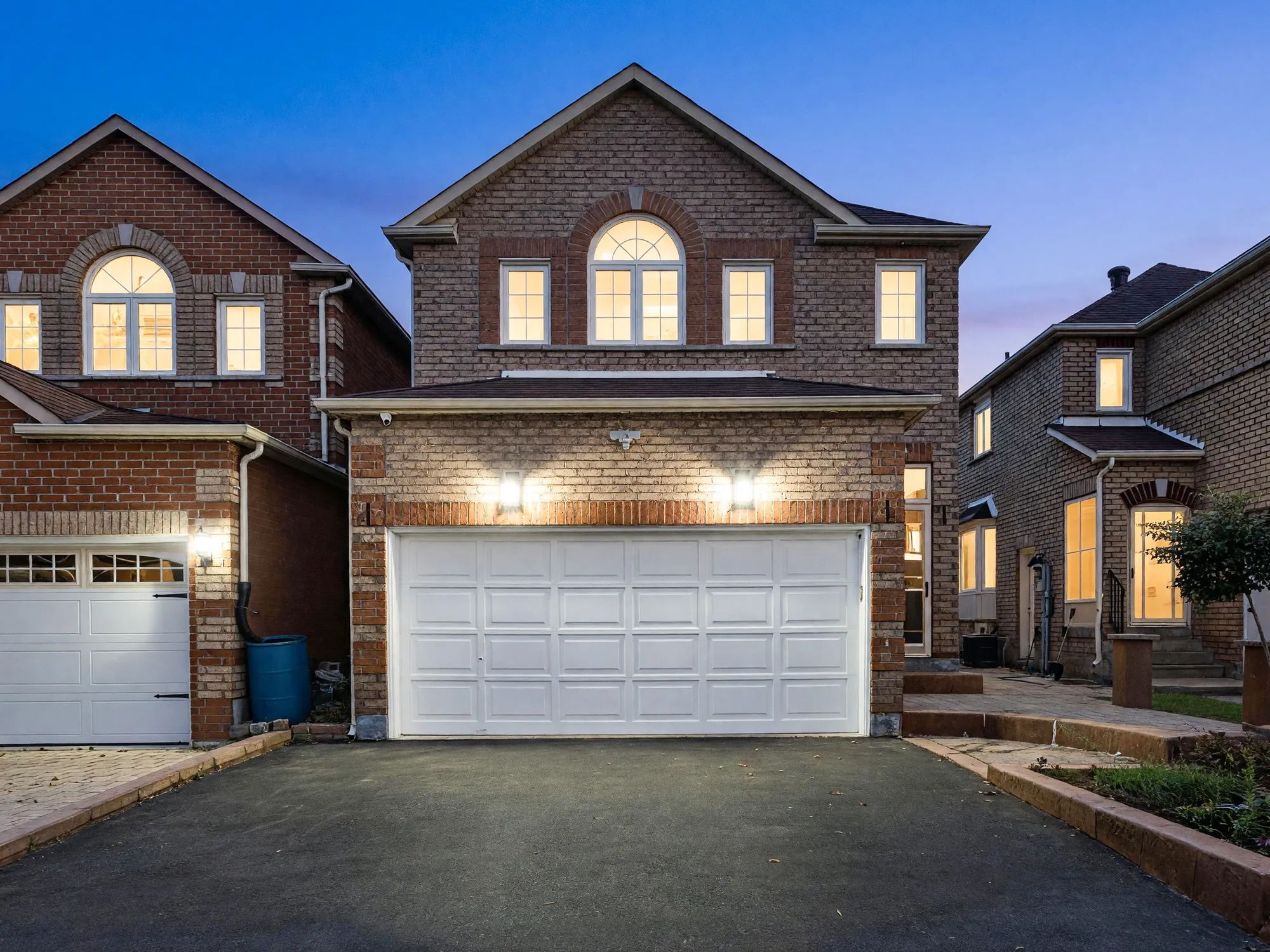 The front of 106 Clarion Crescent at dusk: two storeys of brick under a hip roof, an arched window over the white double garage door, the windows and the garage lanterns lit against a deep blue sky