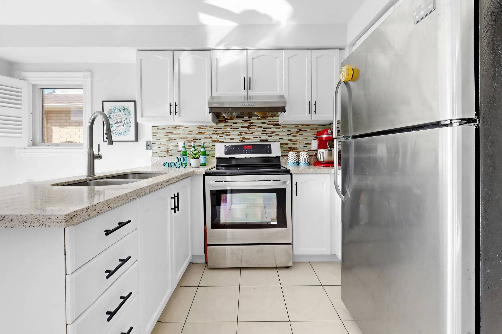 The kitchen: white cabinets, a stainless range and a stainless refrigerator, a mosaic tile backsplash, a window over the double sink