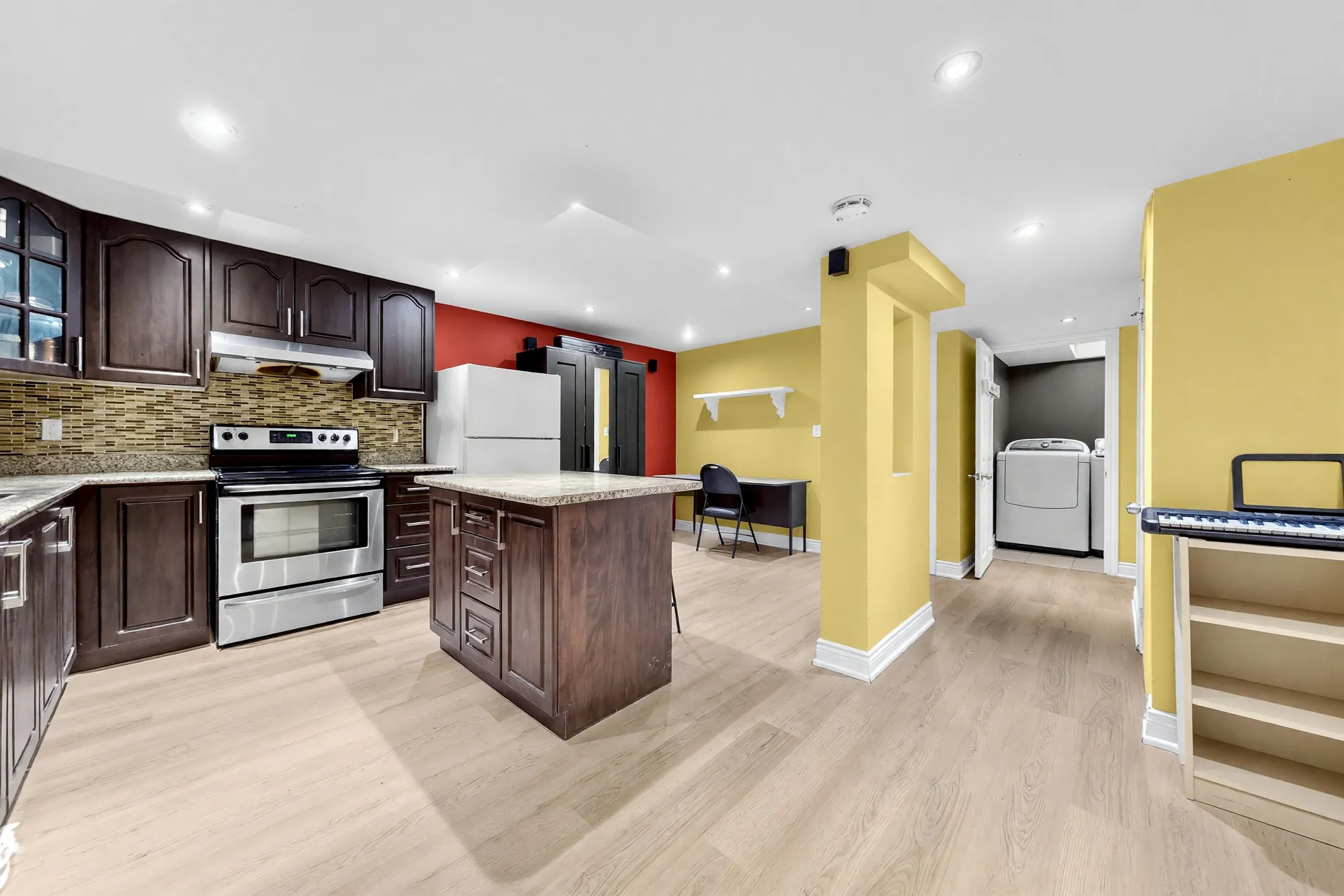 The lower-floor kitchen shown cleared: dark wood cabinets, a stainless range, a mosaic backsplash, an island, a refrigerator by the red wall