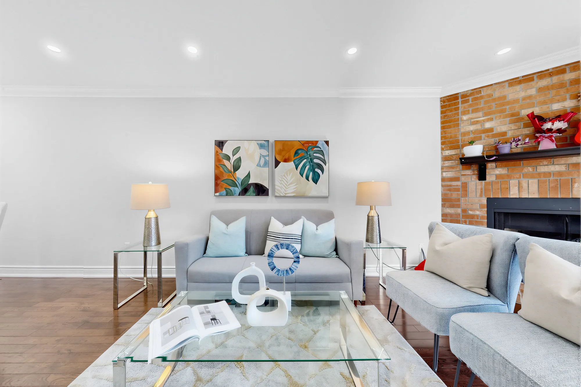 The living room's sofa between two lamps under a pair of leaf prints, the brick fireplace to the right, a glass coffee table