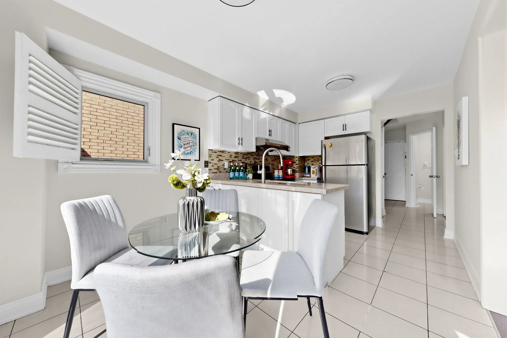 The breakfast nook: a round glass table with four chairs by a window with shutters, the kitchen beyond, tile floor