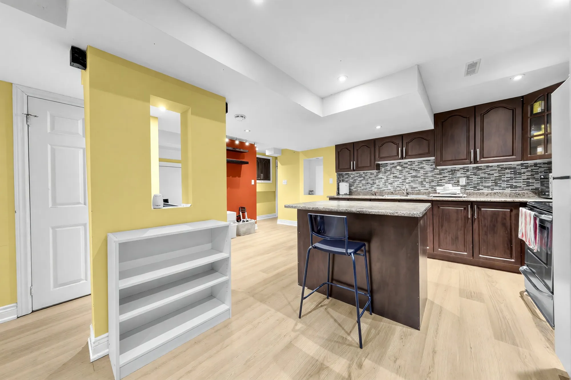 The open office area shown cleared: an empty white shelf unit by the yellow column, the kitchen island with a stool, the cabinets beyond