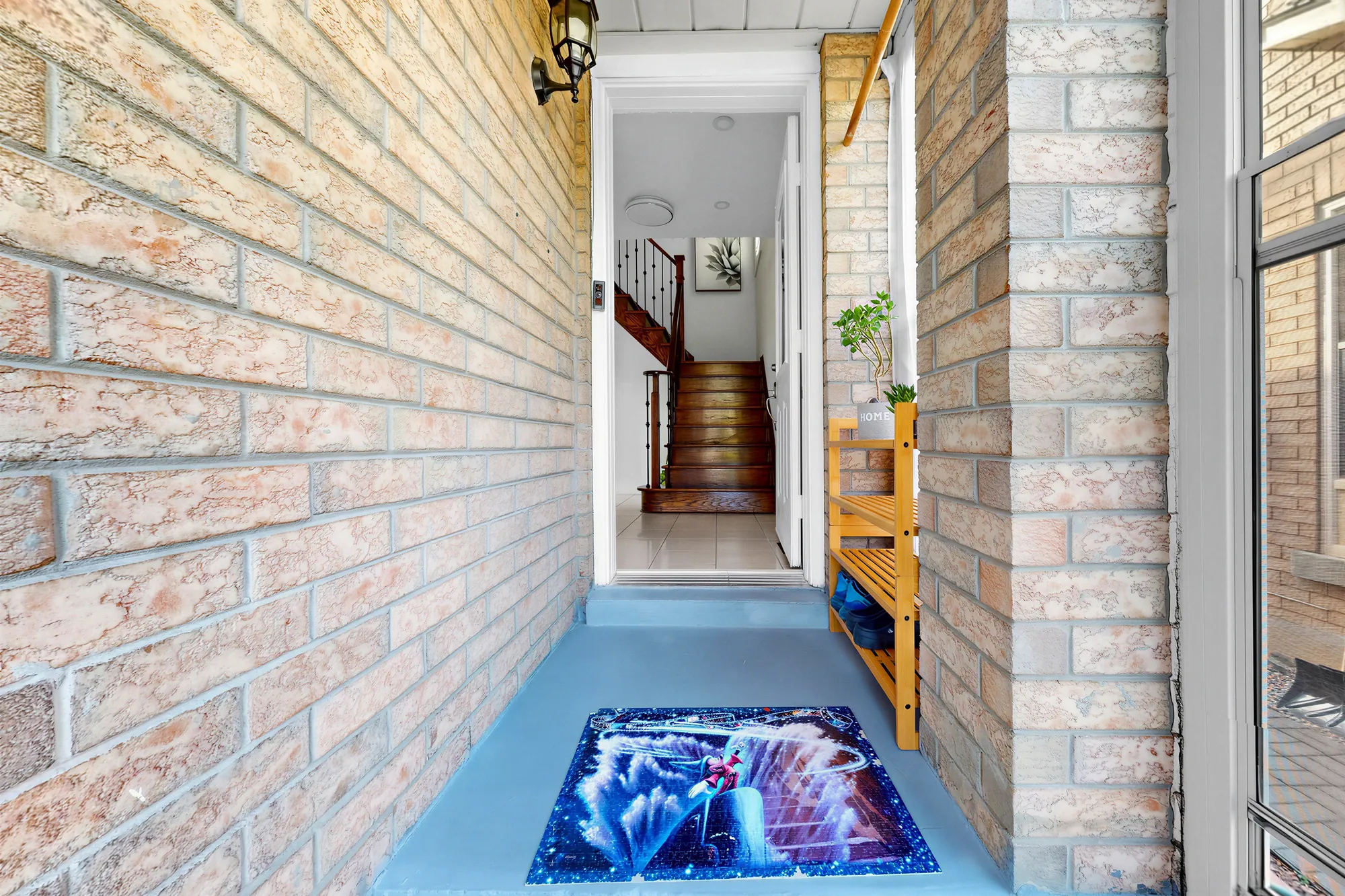 From the porch through the house door into the foyer, the oak stair ahead, a shoe bench along the brick wall