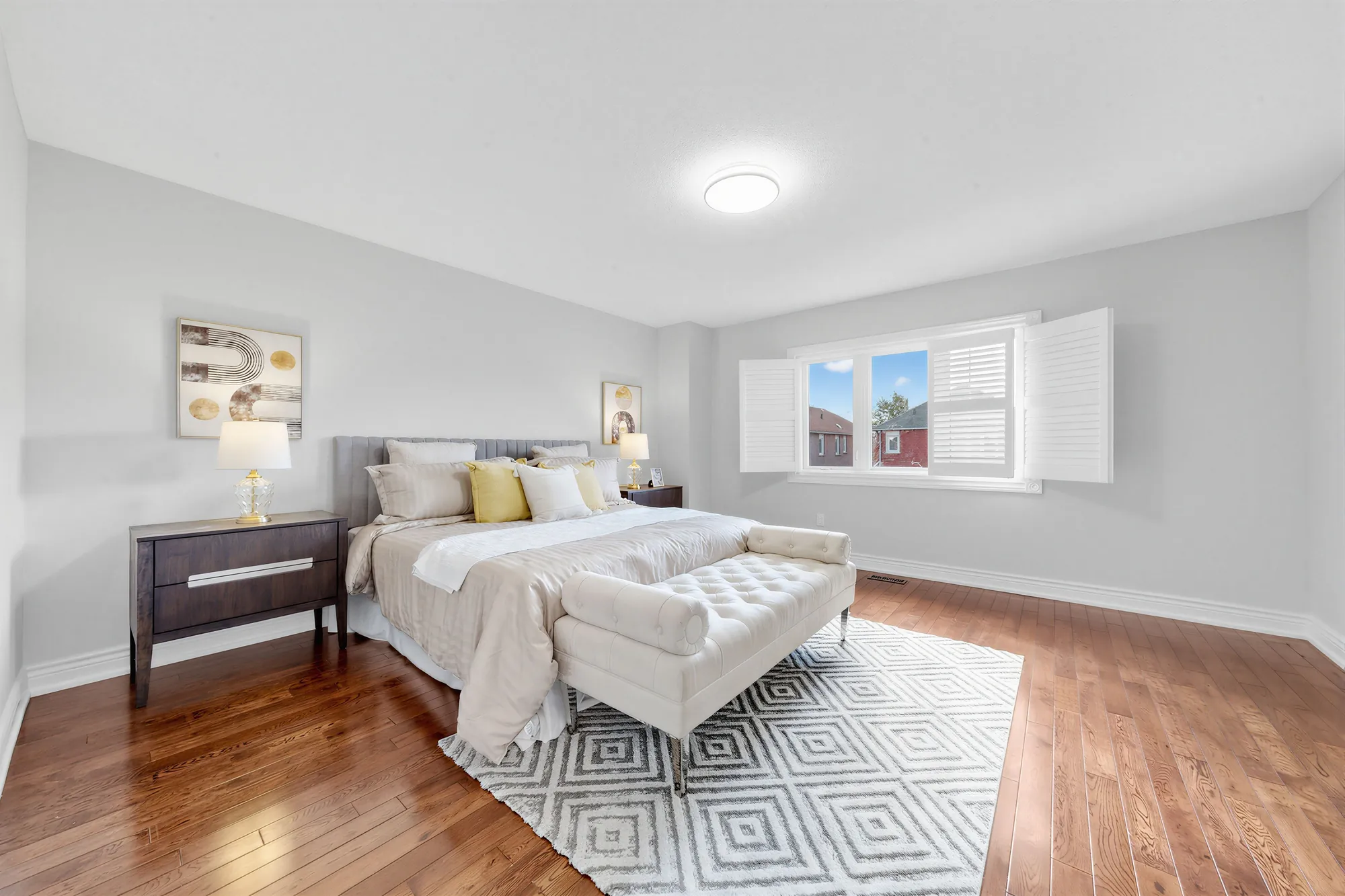 The primary bedroom: hardwood floors, a bed with a tufted bench on a geometric rug, night tables and lamps, a window with white shutters